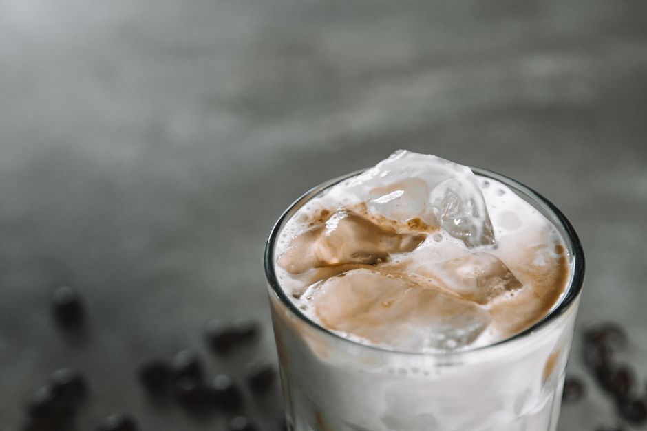 Verre de café glacé crémeux avec des glaçons, sur un fond gris et quelques grains de café en arrière-plan.