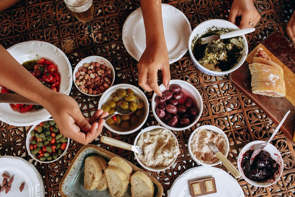 Table garnie de plats variés, incluant houmous, olives, tomates et pain, avec des mains qui se servent.