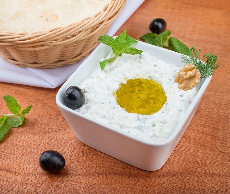 Tzatziki maison dans un bol blanc, garni d'huile d'olive, olives noires et noix, avec un panier de pain en arrière-plan.