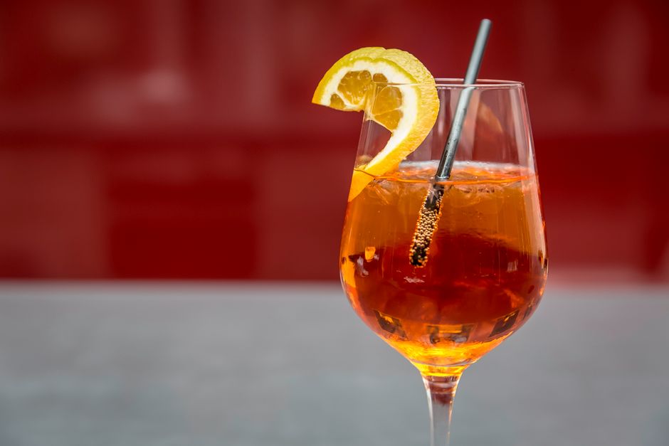 Verre de spritz maison avec une rondelle d'orange et une paille, sur fond rouge.