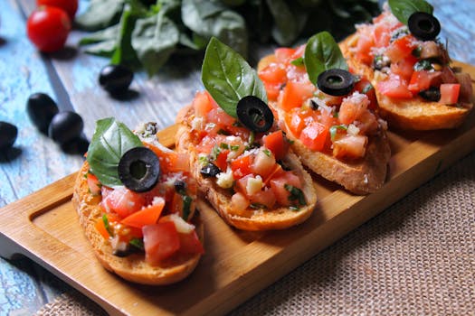 Quatre bruschettas garnies de tomates, olives et basilic sur une planche en bois, avec des ingrédients frais en arrière-plan.