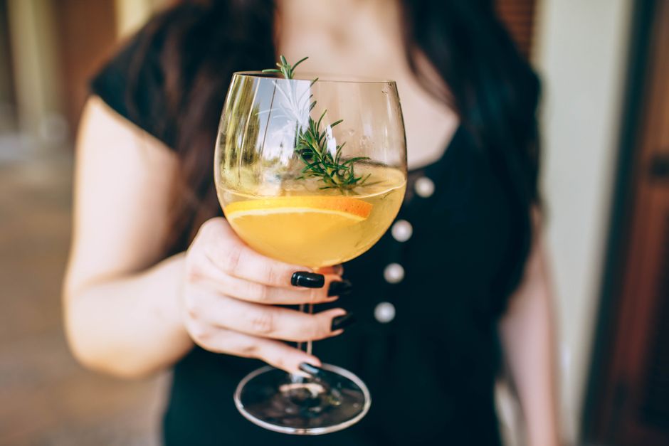 Verre de sangria blanche avec des tranches d'orange et des brins de romarin, tenu par une personne en noir.