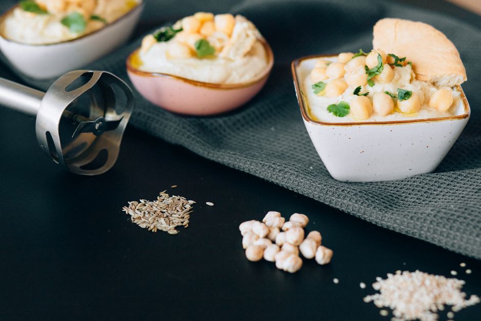 Tartinade de houmous maison servie dans des bols avec des pois chiches et des herbes, accompagnée d'un mixeur et d'épices.