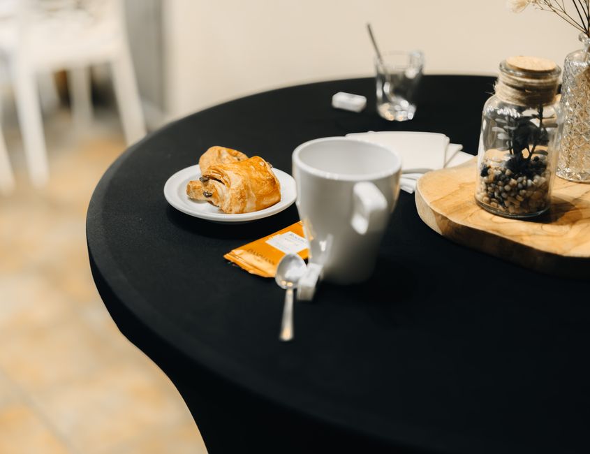 Table noire avec une tasse blanche, un croissant, un sachet de sucre et un pot de fleurs séchées. Ambiance apéritif convivial