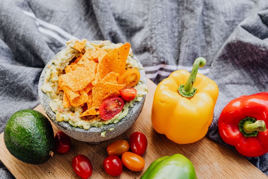 Bol de guacamole garni de chips, tomates cerises et poivrons colorés sur une planche en bois.