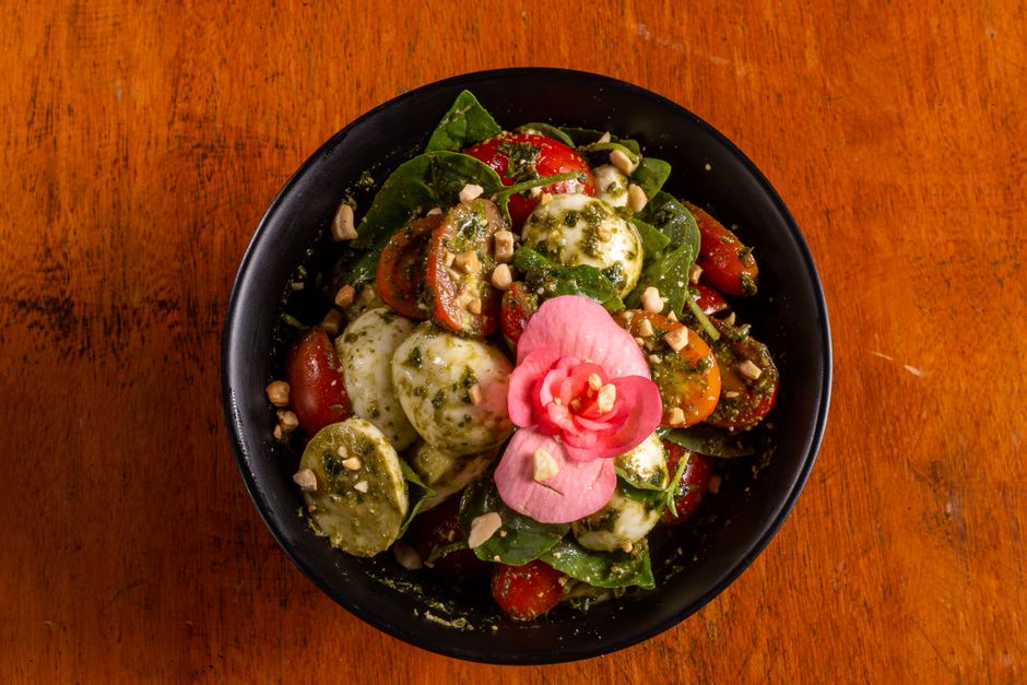 Salade de tomates cerises, mozzarella et basilic, garnie de fleurs comestibles et de noix, servie dans un bol noir.