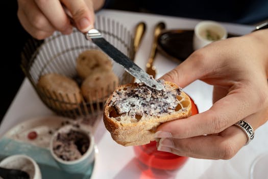 Main étalant de la tapenade sur un morceau de pain, avec des pains en arrière-plan et des couverts élégants.