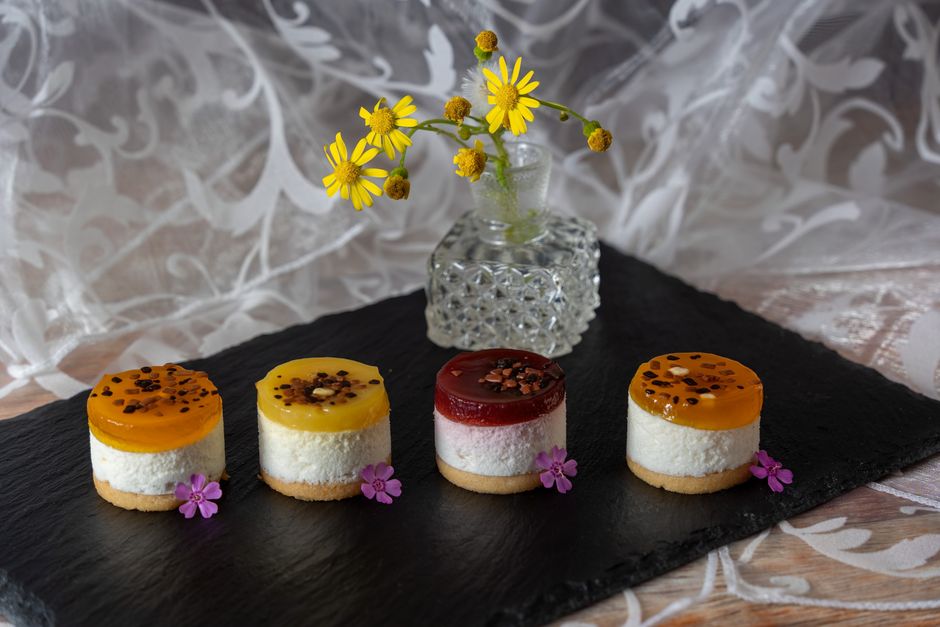 Quatre mini desserts colorés sur un plateau noir, garnis de gelée et de fleurs comestibles, avec un vase de fleurs en arrière