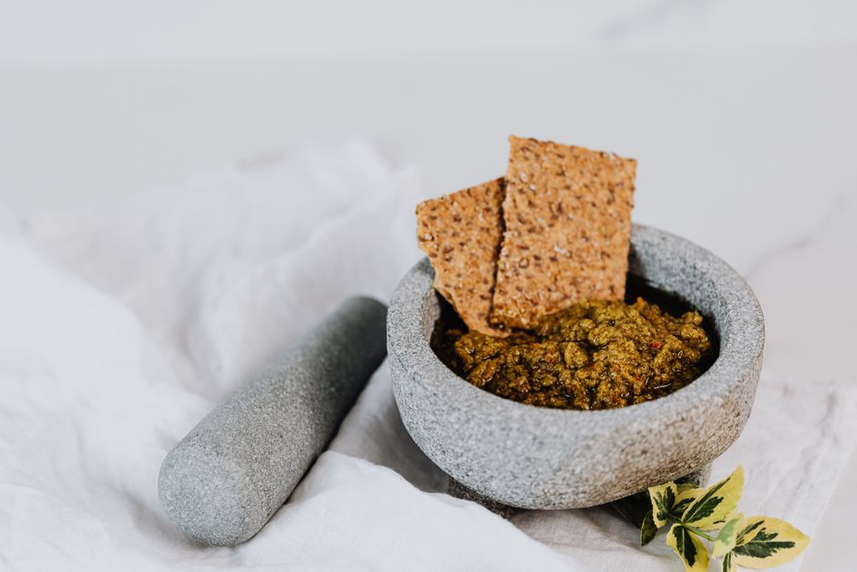 Crackers aux graines posés sur un bol de tartinade, avec un pilon en pierre à côté sur un tissu blanc.