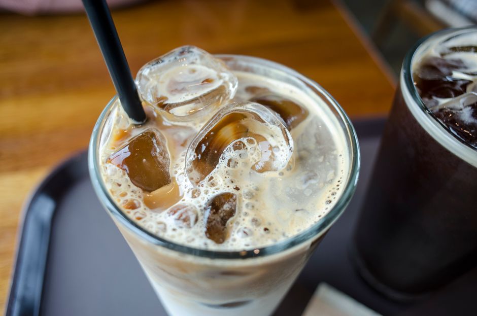 Verre de café glacé crémeux avec des glaçons et une paille, sur un plateau en bois.