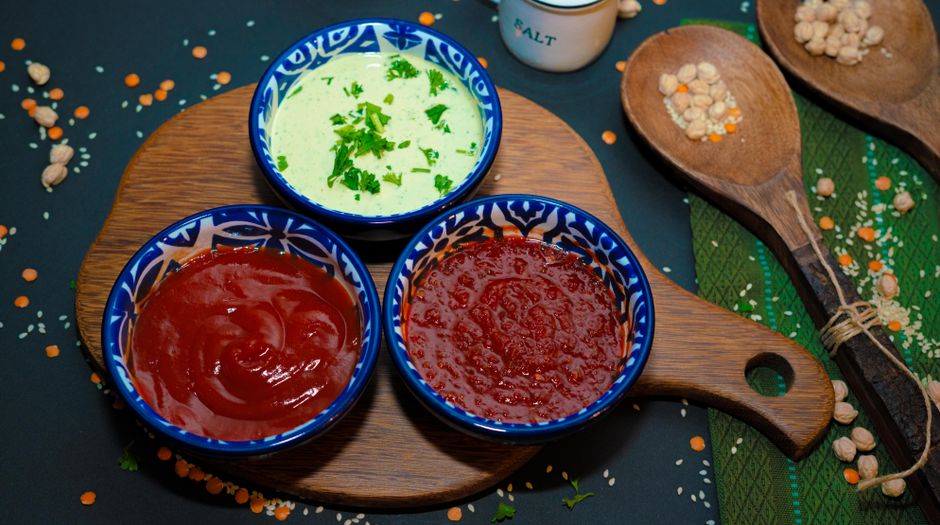 Trois bols de sauces colorées sur une planche en bois, entourés de lentilles et de noix.