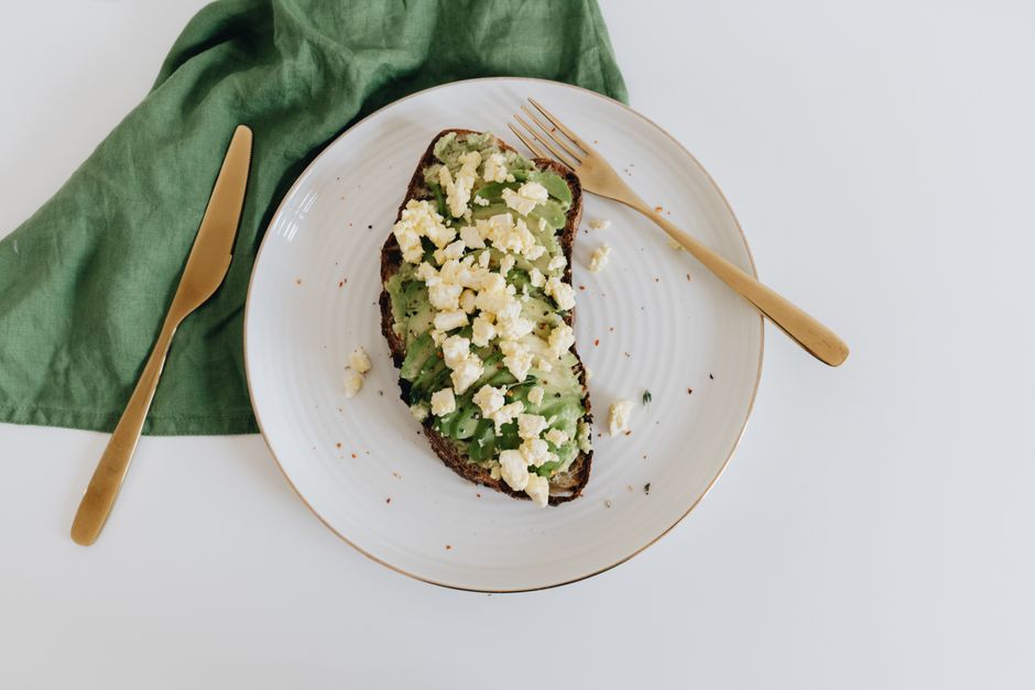 Tartine de pain complet garnie d'avocat et de crème de feta sur une assiette blanche avec des couverts dorés.