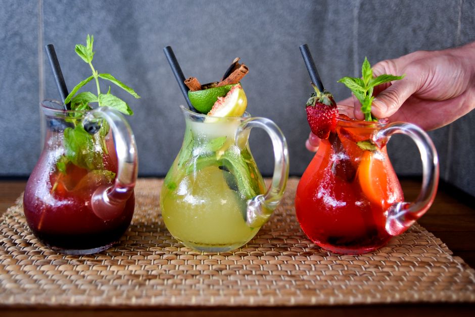Trois carafes de sangria colorées avec fruits et herbes, sur un plateau en osier, sur fond gris.